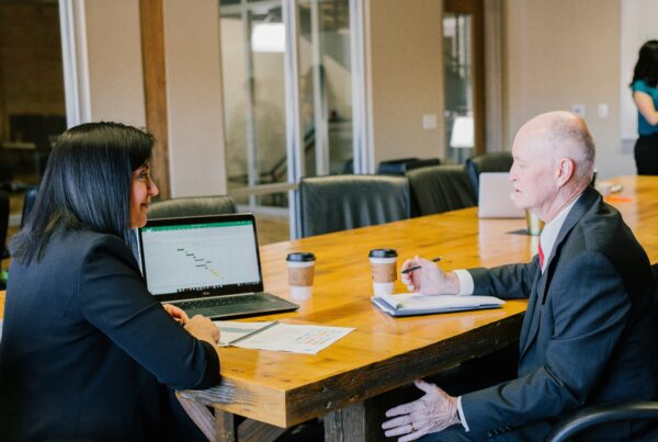 A dark-haired woman shows data on a laptop to an older man. They are sitting at a conference table in a meeting room.
