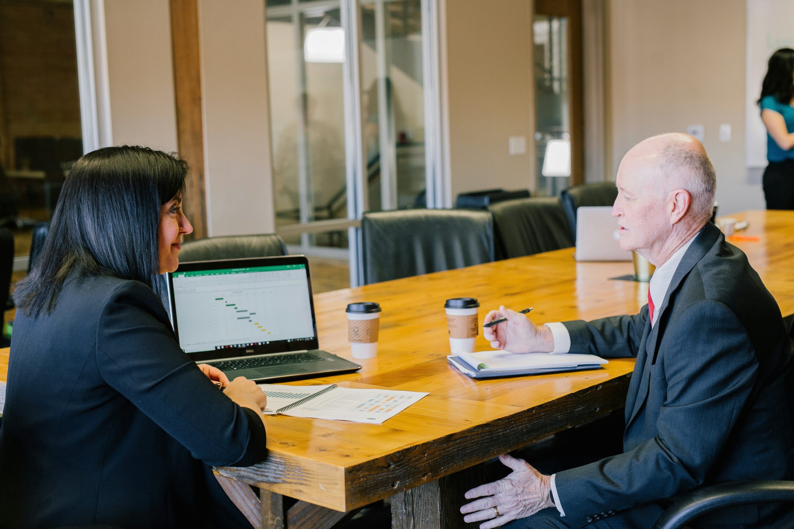 A dark-haired woman shows data on a laptop to an older man. They are sitting at a conference table in a meeting room.